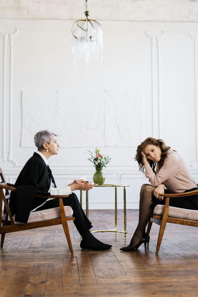 Two women sitting in wooden chairs facing each other in a bright room, with one appearing to listen attentively and the other resting her head on her hand.