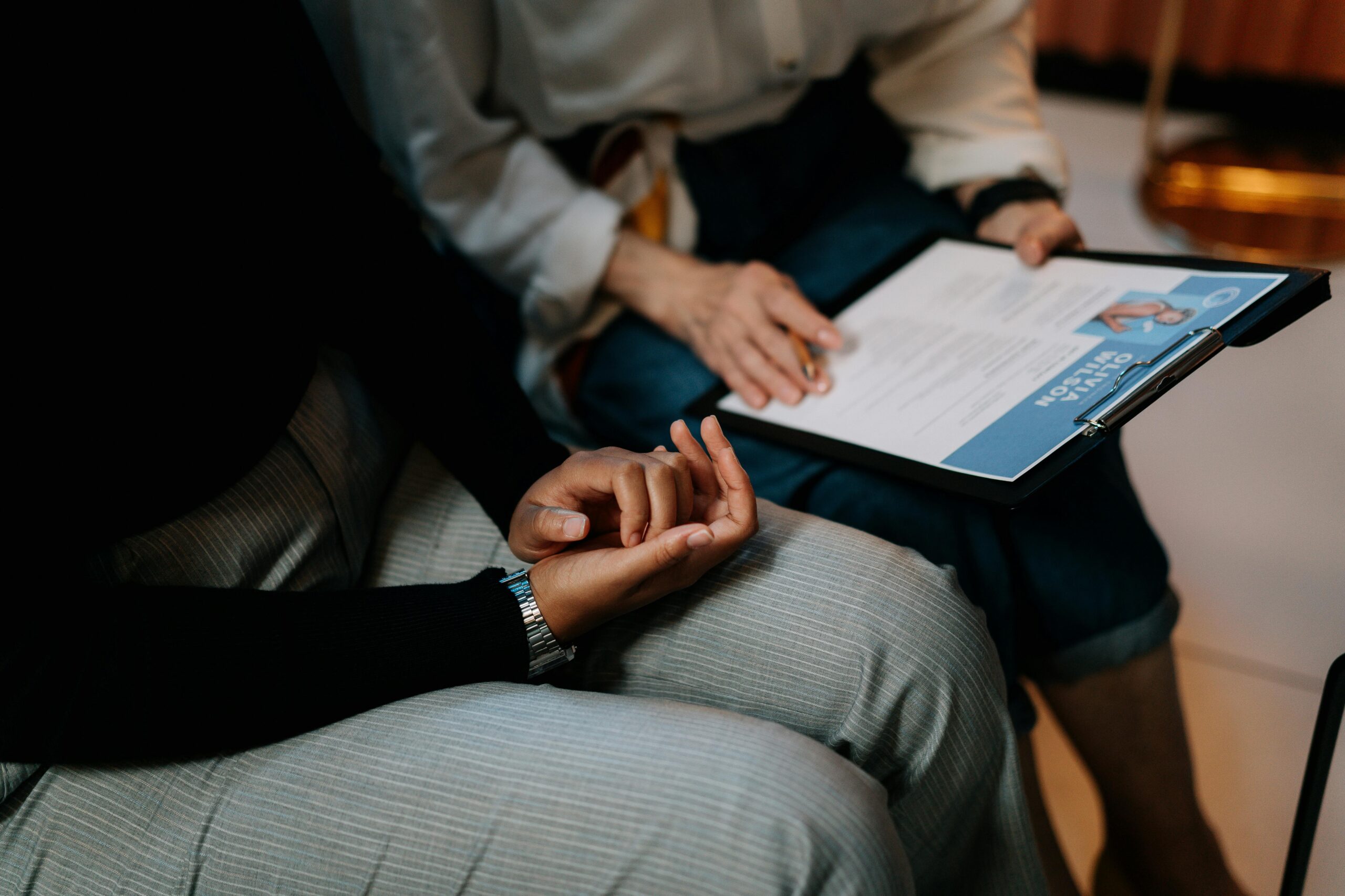 Two people sitting close together, with one person holding a clipboard and paper while the other clasps their hands.