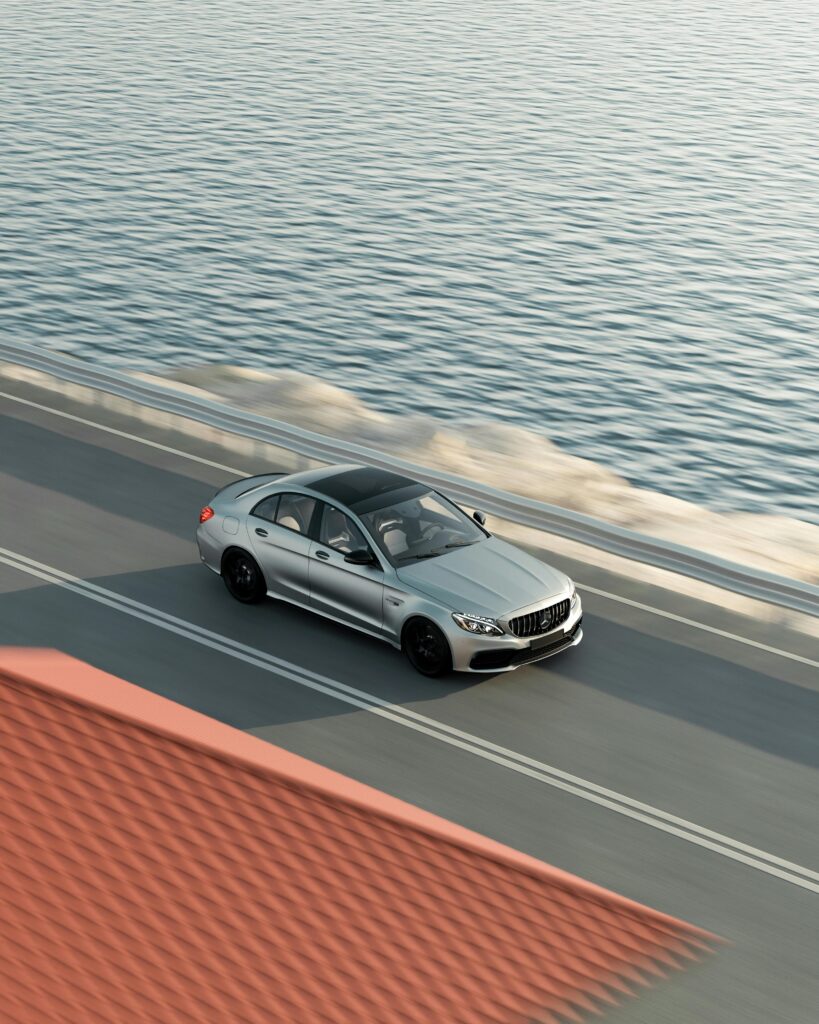 **Alt text:** A silver car drives along a coastal road with a body of water on one side and a building with a red roof on the other.