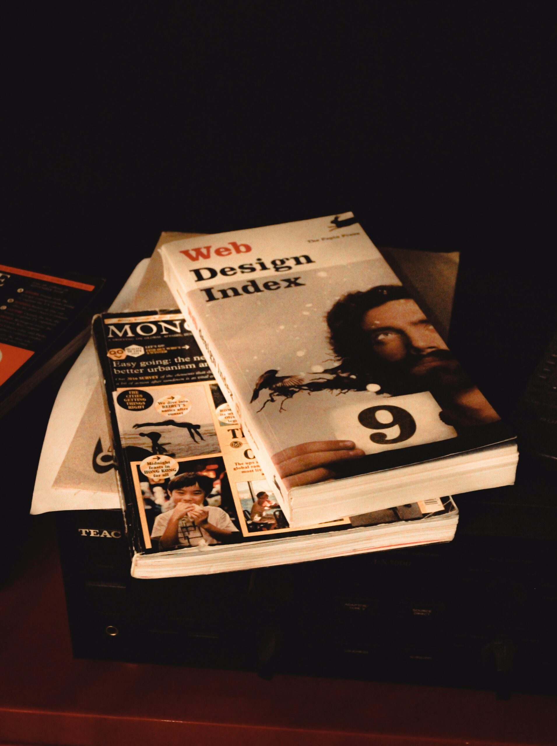 **Alt Text:** A stack of books and magazines, including one titled "Web Design Index," placed on a dark surface.