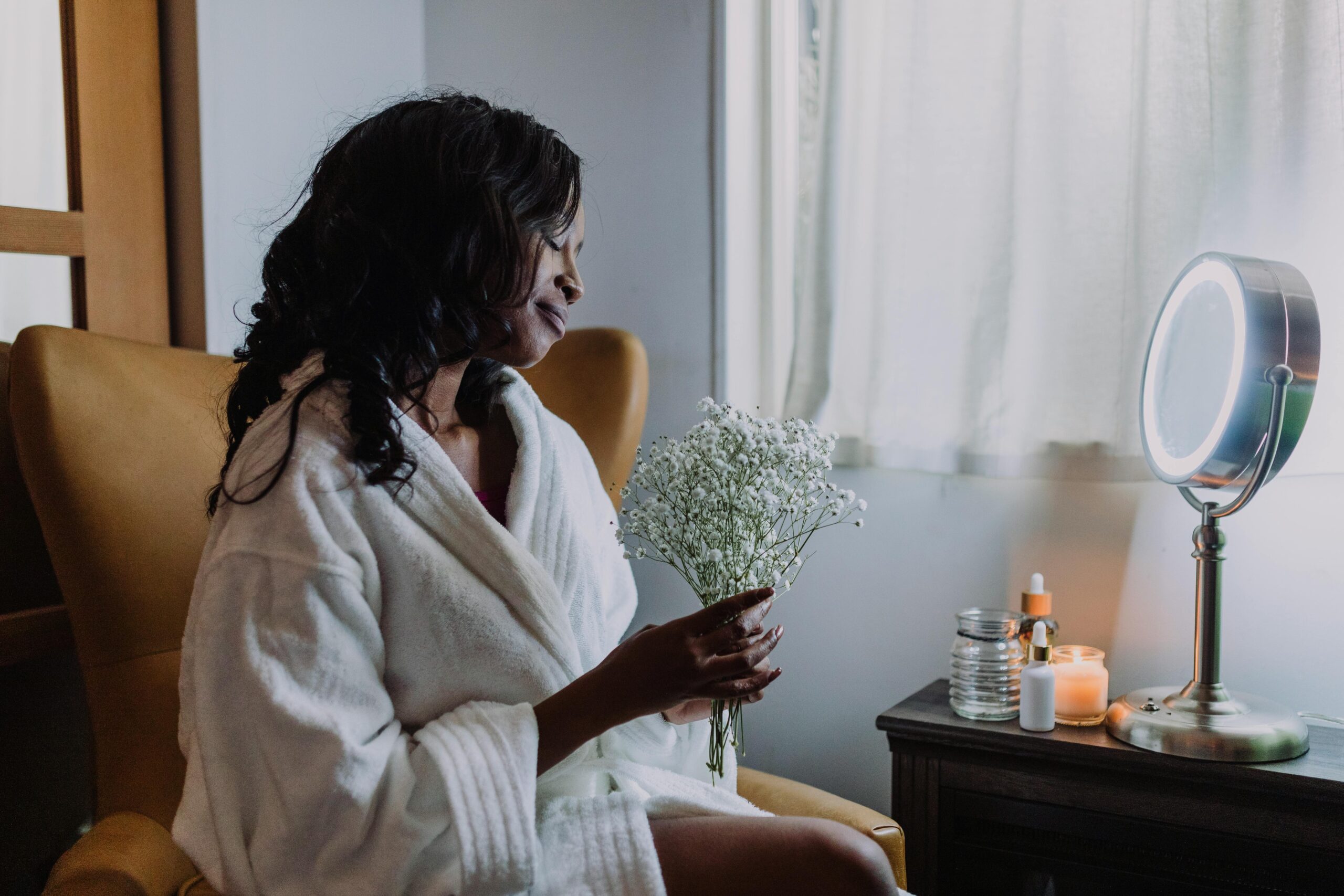 A woman in a white robe sits in a chair holding a bouquet of small white flowers, next to a lit makeup mirror and candles on a table.