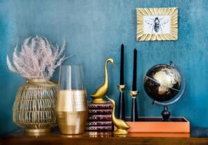 A stylish shelf decorated with gold accents, including a globe, candle holders, a swan figurine, and a framed bee illustration, set against a blue background.