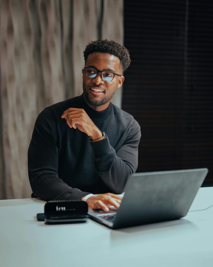 A man wearing glasses and a black sweater is sitting at a desk with a laptop, looking to the side and smiling.