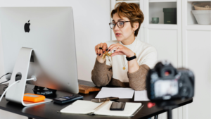 A woman in glasses sits at a desk with a computer, papers, and a camera, gesturing with her hands as if speaking or presenting.