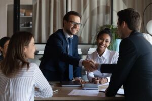 Four people in a business meeting, with two men shaking hands while two women look on and smile.