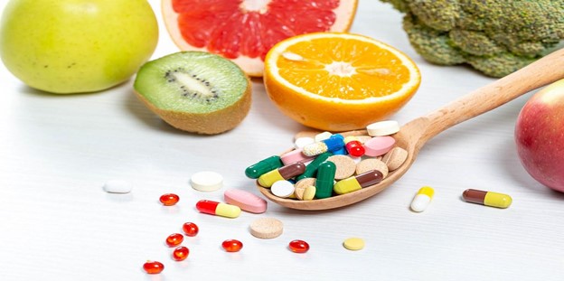 **Alt Text:** A wooden spoon filled with various pills and supplements surrounded by fruits like apple, kiwi, grapefruit, orange, and broccoli.