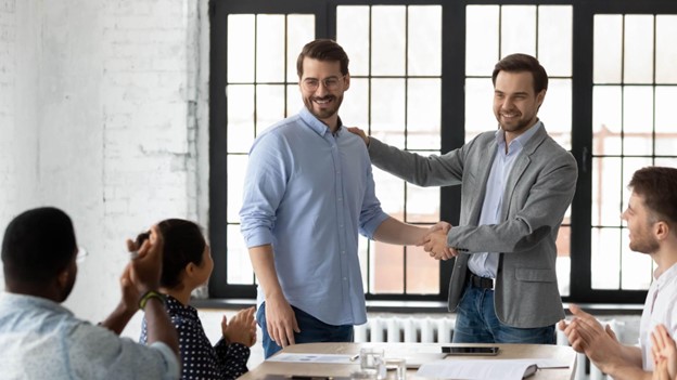 **Alt Text:** Two men stand in front of a window, one shaking hands while the other pats his shoulder, as three seated individuals applaud.