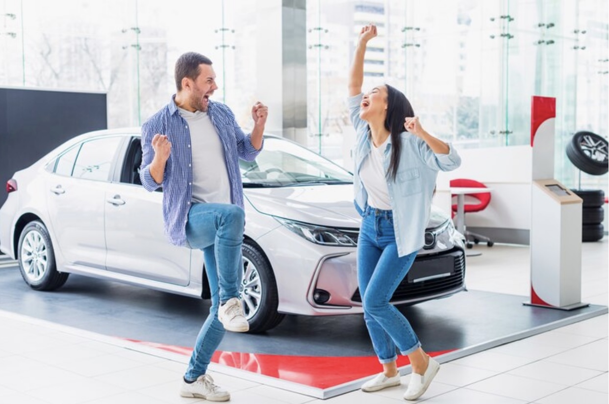 **Alt Text:** A man and a woman joyfully celebrate in front of a silver car inside a showroom.