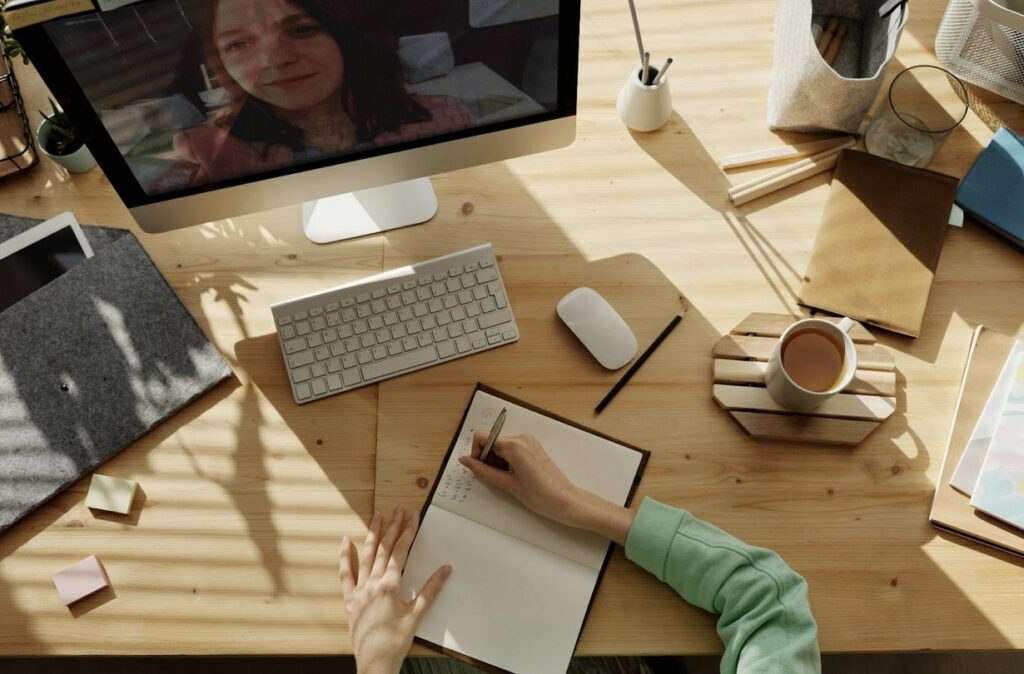 A person writing in a notebook on a wooden desk with a computer showing a video call, surrounded by office supplies and a cup of coffee.