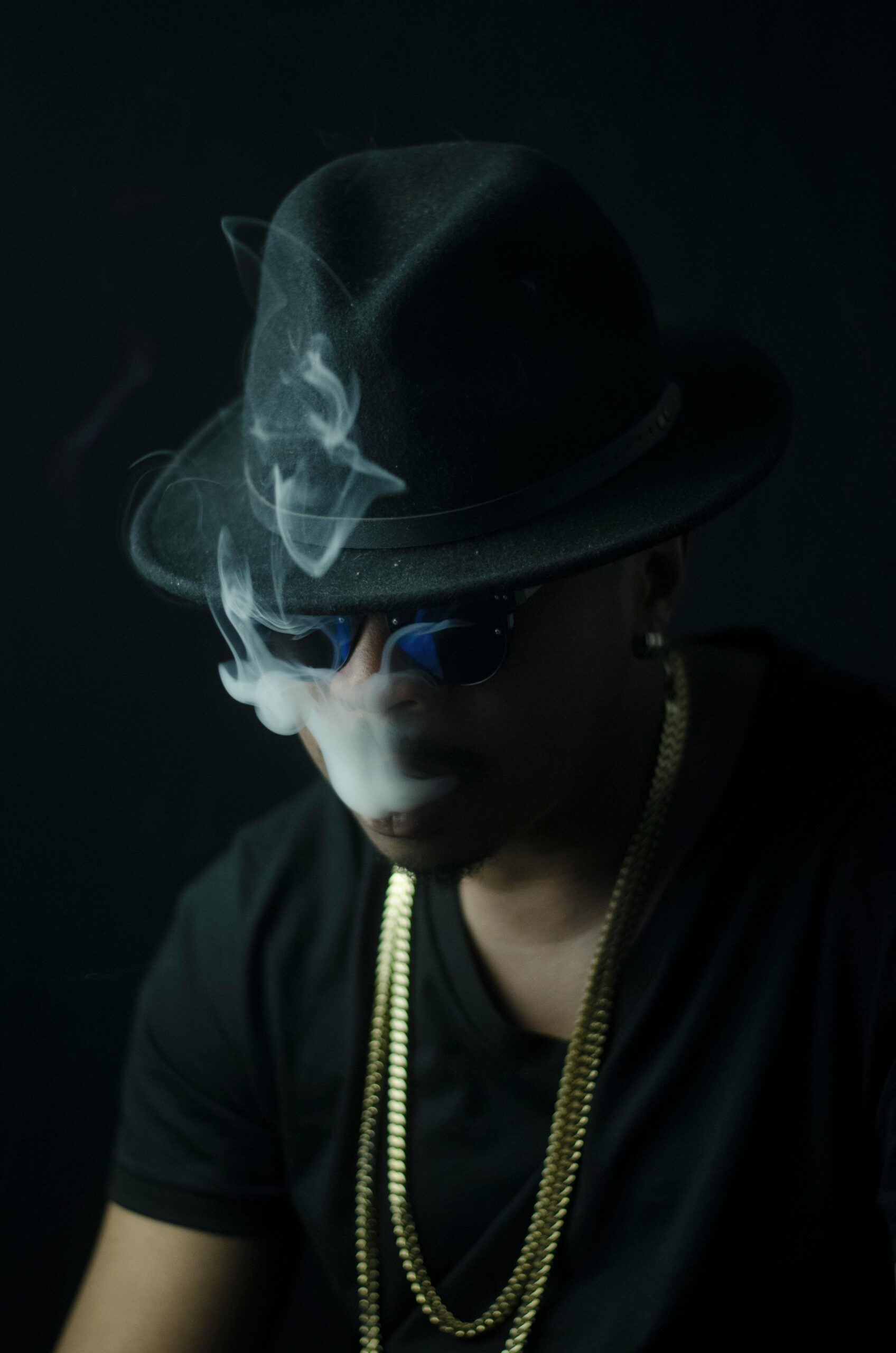 A person wearing a hat, sunglasses, and gold chains exhales smoke in a dimly lit setting.