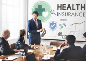 **Alt Text:** A group of business professionals in a meeting, with a man in a suit presenting healthcare insurance concepts displayed on a screen behind him.