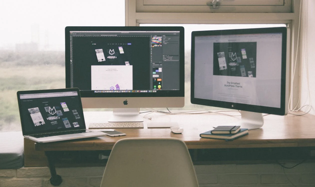 A workspace with a laptop and two large desktop monitors displaying creative design software and webpage layouts, situated by a window.