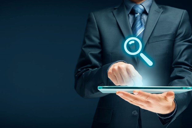 A person in a suit uses a digital tablet with a glowing magnifying glass icon appearing above it.