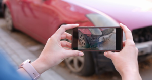 A person is taking a photo of a damaged red car's front bumper with a smartphone.
