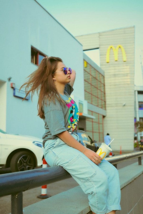 **Alt text:** A person wearing sunglasses and a graphic t-shirt sits on a railing outside a McDonald's, holding a drink cup.