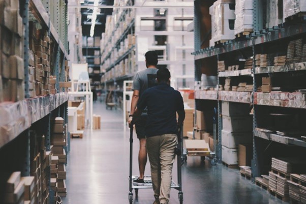 **Alt Text:** Two people navigate through a large warehouse with tall shelves filled with boxes, one pushing the other on a cart.