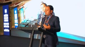 **Alt text:** A man in a suit is speaking at a podium with a microphone in front of a large screen displaying his image.