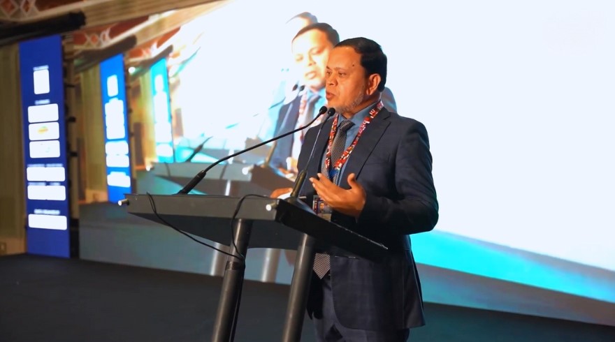 **Alt text:** A man in a suit is speaking at a podium with a microphone in front of a large screen displaying his image.