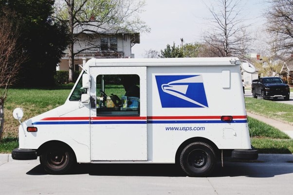 **Alt text:** A USPS mail delivery truck parked on a suburban street with a house and trees in the background.
