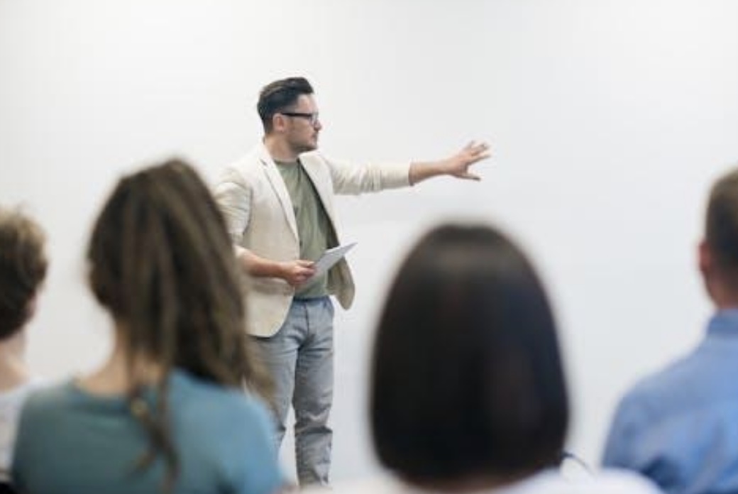 A man in a blazer is gesturing while speaking in front of an audience in a classroom setting.