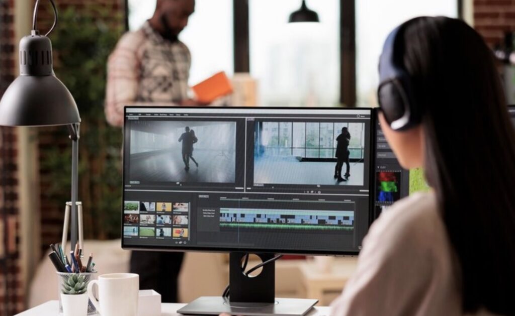 A person wearing headphones is working on video editing software on a computer screen, with another person reading a book in the background.