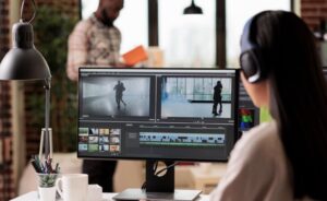 A person wearing headphones is working on video editing software on a computer screen, with another person reading a book in the background.