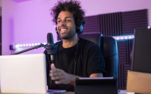 **Alt text:** A person wearing headphones sits in a soundproofed room, speaking into a microphone with a laptop in front.