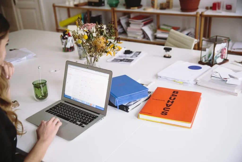A person working on a laptop at a cluttered desk with books, papers, and a vase of flowers.