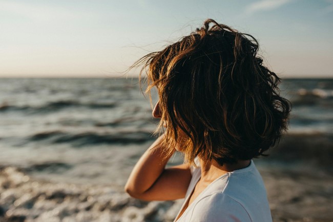 **Alt text**: A person with short hair stands by the ocean, looking towards the horizon under a clear sky.