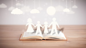 **Alt Text:** A paper cutout of a family of four holding hands is standing on top of an open book, with clouds and a sun hanging above in the background.