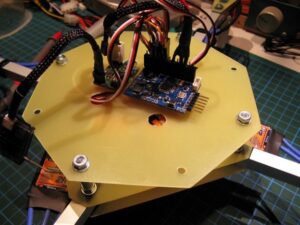 **Alt text:** A circuit board mounted on a hexagonal frame with wires connected, surrounded by other electronic components on a workspace.