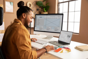 A person sitting at a desk working on a computer, surrounded by color swatches and design layouts on the screen.