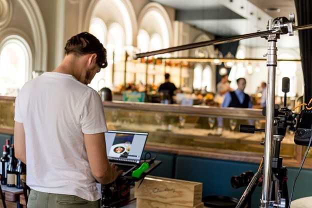 A person in a white t-shirt stands in a restaurant setting, working on a laptop, surrounded by camera equipment, with a busy dining area in the background.