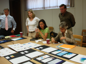 **Alt Text:** A group of five people are gathered around a conference table covered with various documents and charts, engaged in a discussion.