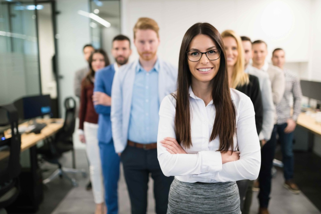 A confident woman in glasses stands at the front of a line of diverse, casually dressed office colleagues in a modern workspace.
