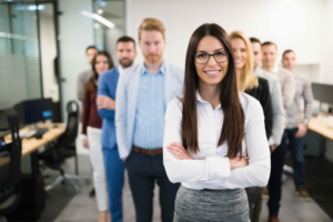 A confident woman in glasses stands at the front of a line of diverse, casually dressed office colleagues in a modern workspace.