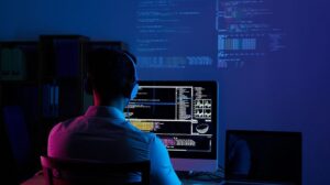 **Alt text:** A person wearing headphones sits in front of computer screens displaying complex code and data visualizations in a dimly lit room.