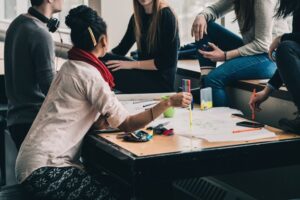 **Alt text:** A group of people sitting around a table, engaging in discussion with papers and markers spread out.