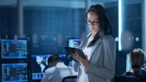 A woman in a business suit uses a tablet in a control room with multiple computer screens showing data and maps.