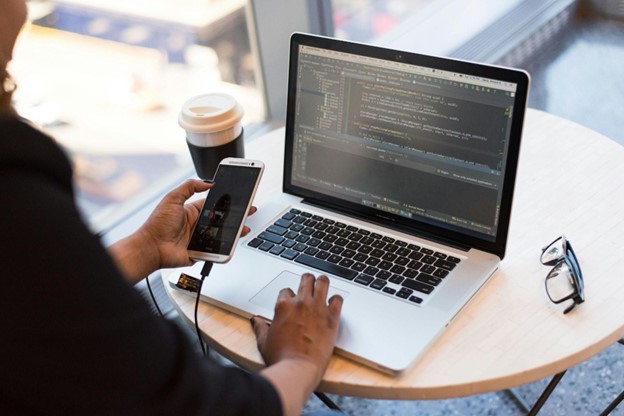 **Alt text:** A person holding a smartphone connected to a laptop displaying programming code, with a coffee cup on the table.