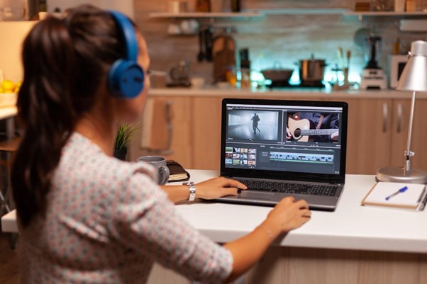 A person wearing blue headphones works on video editing software on a laptop in a kitchen setting.