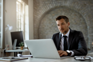 A man in a suit working on a laptop at a desk with a brick archway in the background.