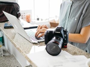 **Alt text:** A person typing on a laptop at a kitchen counter with a professional camera placed nearby.