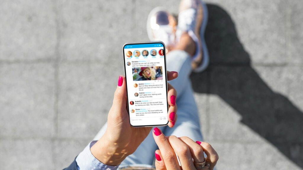 **Alt Text:** A person with bright pink nail polish is looking at a social media feed on a smartphone while sitting cross-legged on a concrete surface.