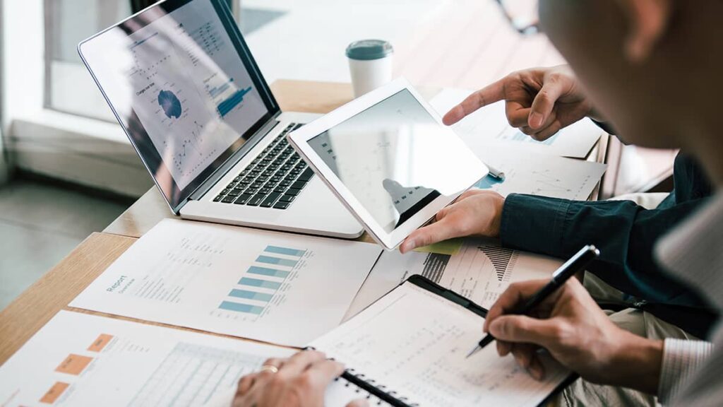 Two people in a meeting are analyzing financial reports on a laptop and tablet, with printed charts on a table.