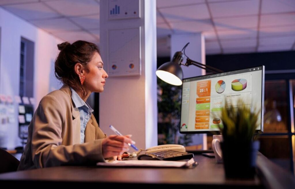 A woman sits in front of a computer displaying charts and graphs, working at a desk under a lamp in a dimly lit office.