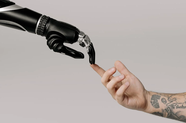 A robotic hand and a human hand with tattoos reaching towards each other, almost touching fingers.