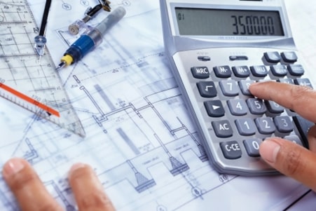 A person using a calculator on top of architectural blueprints with a ruler and pen nearby.