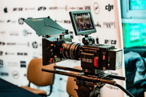 A professional RED digital cinema camera with an external monitor set up on a tripod, with a branded backdrop in the background.
