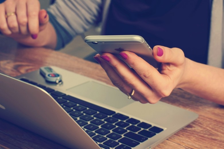 **Alt Text:** A person with pink nail polish holds a smartphone in one hand while resting the other on an open laptop with a USB drive inserted.
